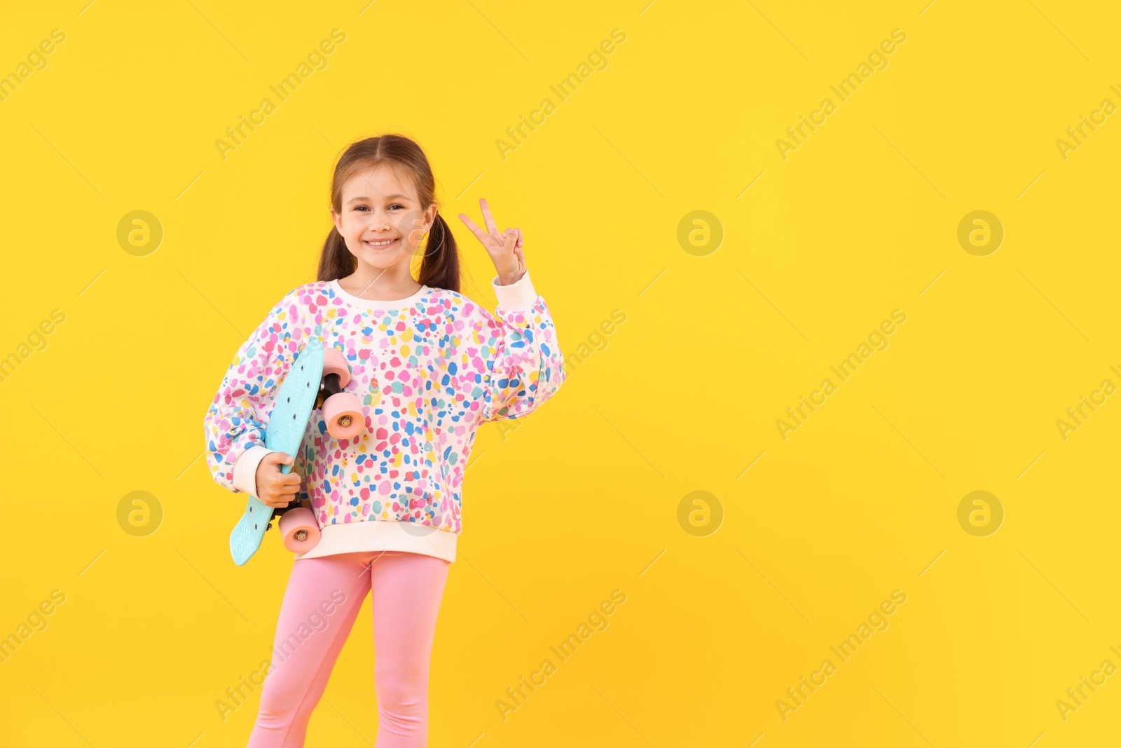 Little girl with penny board showing V sign on yellow background, space for text Photo of Little girl with penny board showing V sign on yellow background, space for text