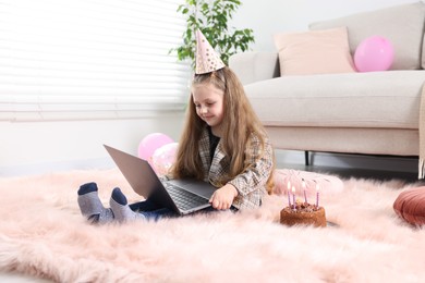 Photo of Birthday celebration. Little girl with cake having video call via laptop at home