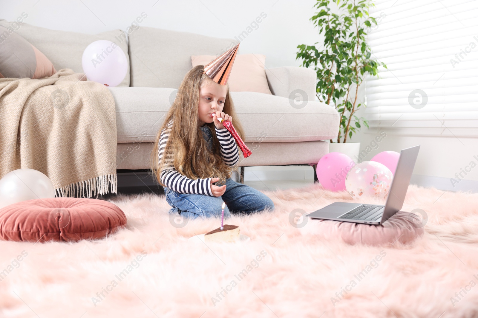 Photo of Birthday celebration. Little girl with blower and piece of cake having video call via laptop at home