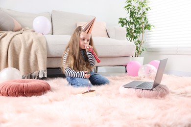 Photo of Birthday celebration. Little girl with blower and piece of cake having video call via laptop at home
