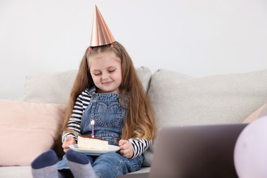 Photo of Birthday celebration. Little girl with piece of cake having video call via laptop on sofa at home