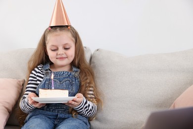 Photo of Birthday celebration. Little girl with piece of cake having video call via laptop on sofa at home