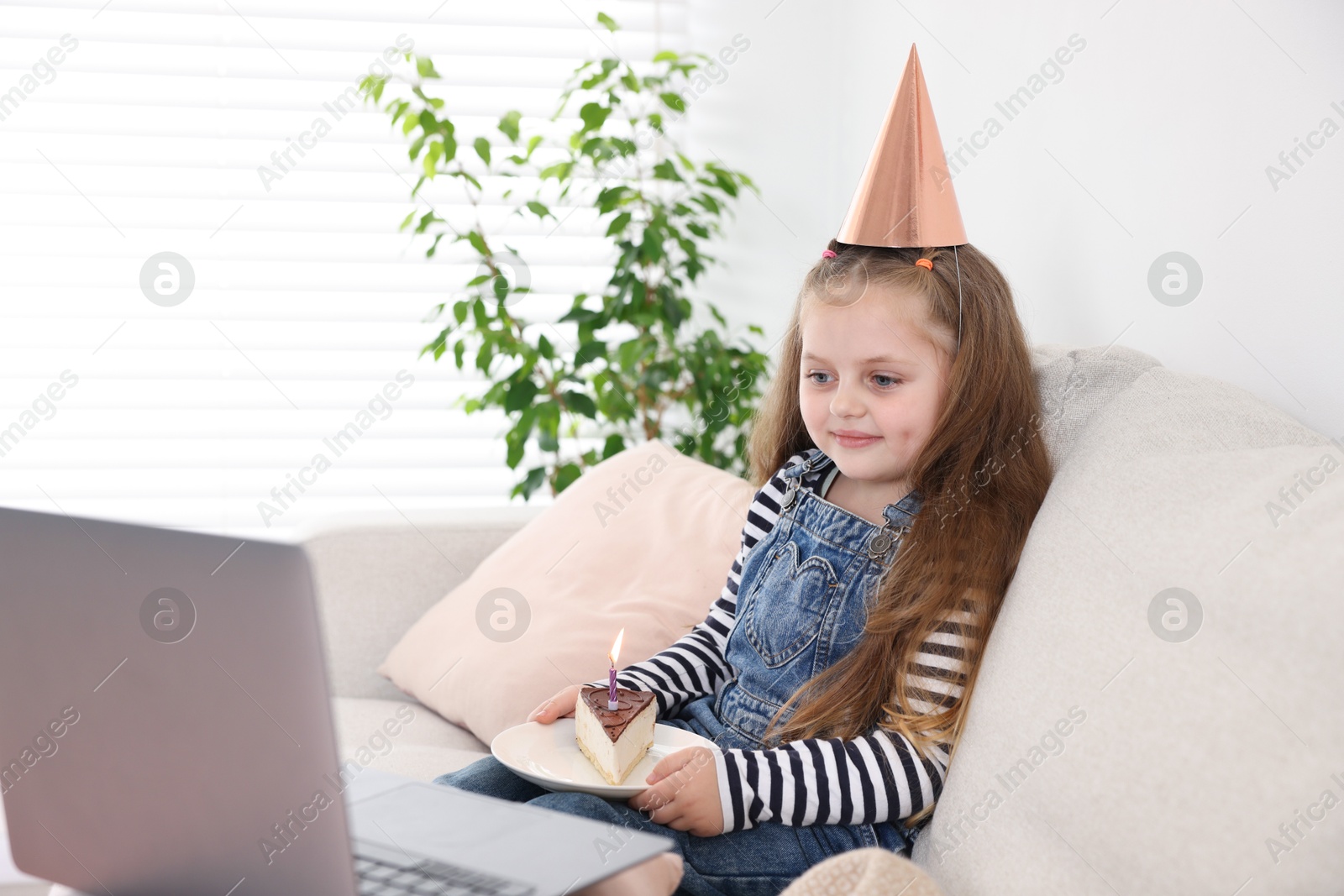 Photo of Birthday celebration. Little girl with piece of cake having video call via laptop on sofa at home