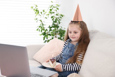 Photo of Birthday celebration. Little girl with piece of cake having video call via laptop on sofa at home
