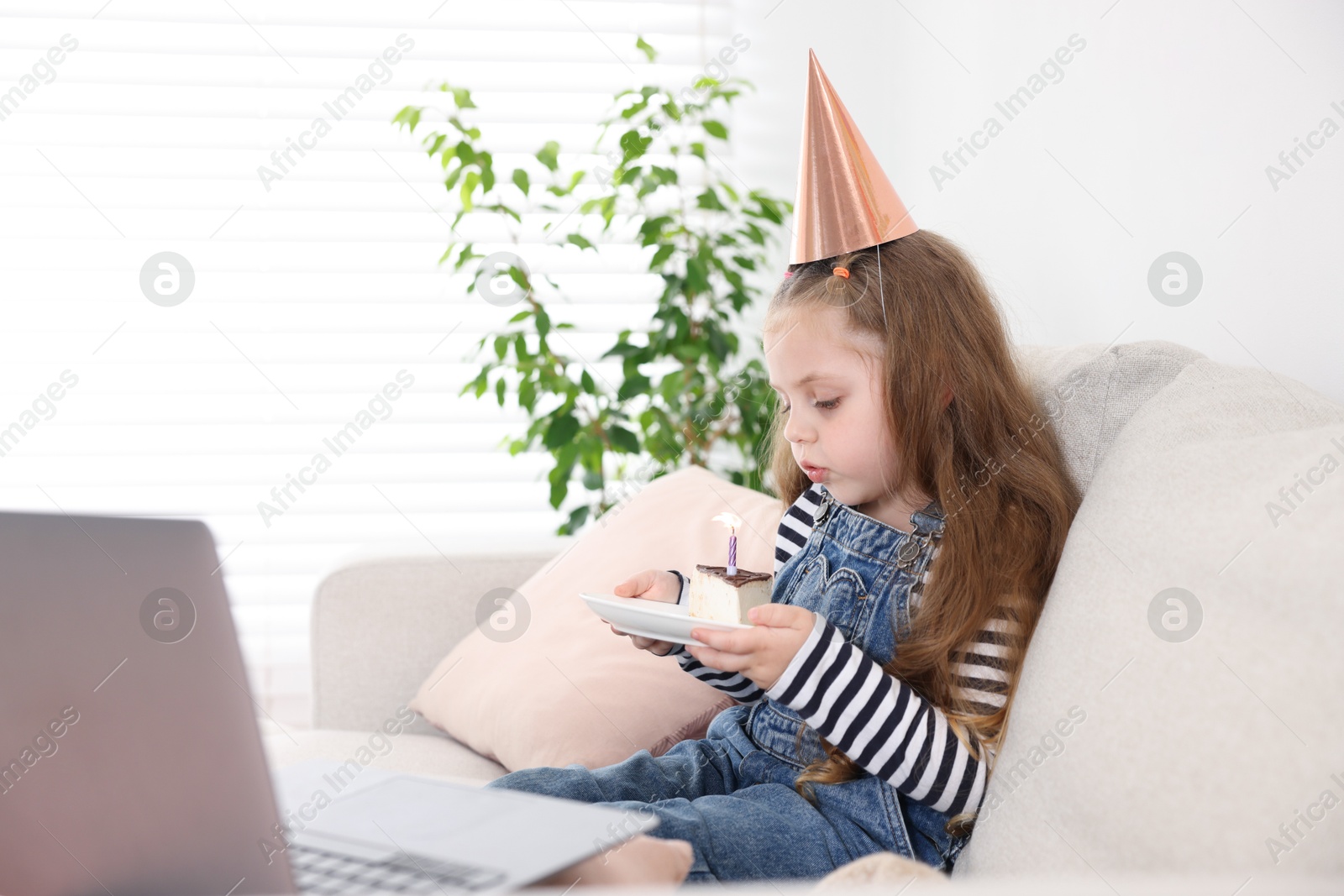 Photo of Birthday celebration. Little girl blowing candle on piece of cake while having video call via laptop on sofa at home