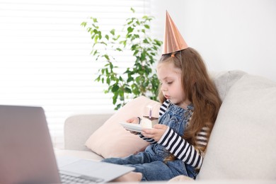 Photo of Birthday celebration. Little girl blowing candle on piece of cake while having video call via laptop on sofa at home