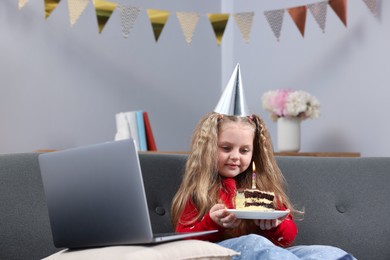 Photo of Birthday celebration. Little girl with piece of cake having video call via laptop on sofa at home