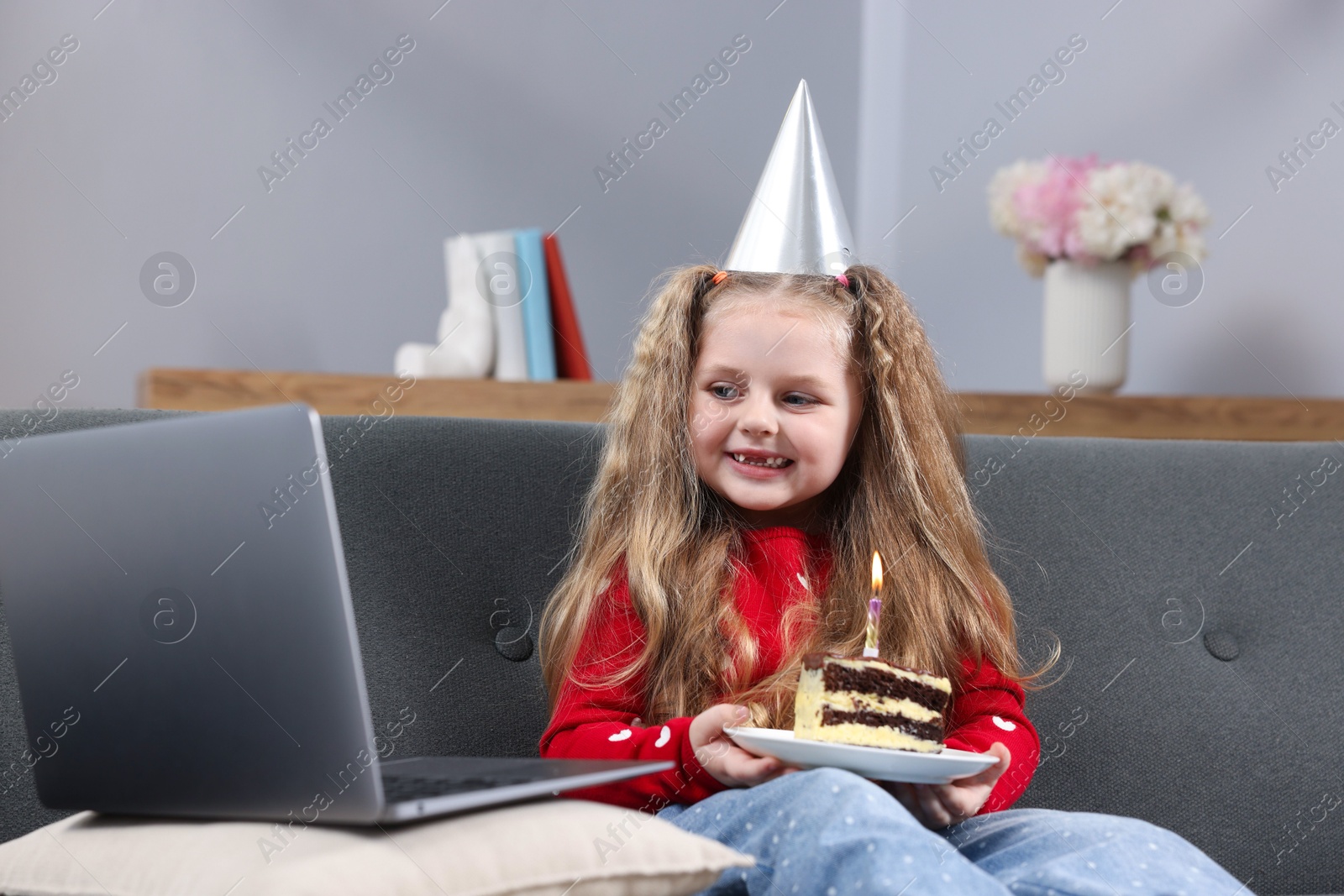 Photo of Birthday celebration. Little girl with piece of cake having video call via laptop on sofa at home