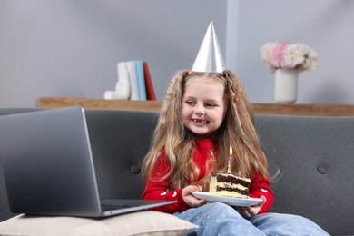 Photo of Birthday celebration. Little girl with piece of cake having video call via laptop on sofa at home