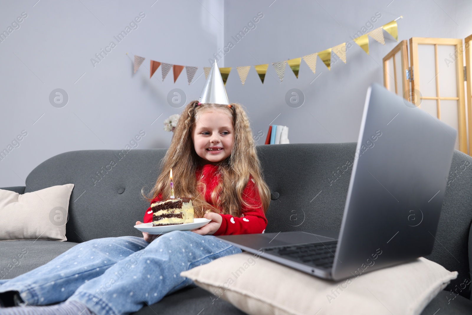 Photo of Birthday celebration. Little girl with piece of cake having video call via laptop on sofa at home