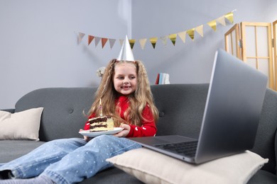 Photo of Birthday celebration. Little girl with piece of cake having video call via laptop on sofa at home