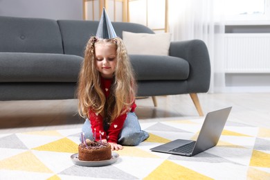 Photo of Birthday celebration. Little girl with cake having video call via laptop at home