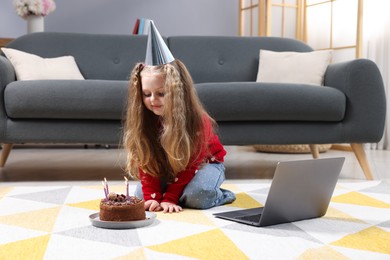Photo of Birthday celebration. Little girl with cake having video call via laptop at home