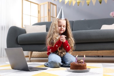 Photo of Birthday celebration. Little girl with cake having video call via laptop at home