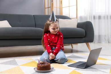 Photo of Birthday celebration. Little girl with cake having video call via laptop at home