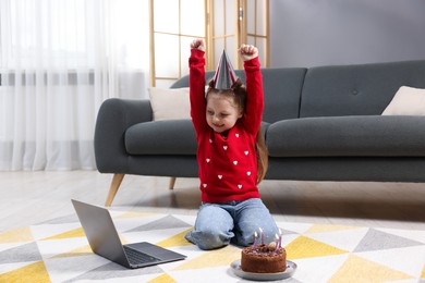 Photo of Birthday celebration. Little girl with cake having video call via laptop at home