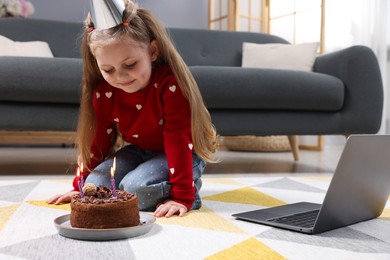 Photo of Birthday celebration. Little girl with cake having video call via laptop at home