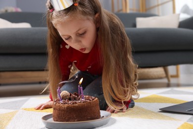 Photo of Birthday celebration. Little girl blowing candles on cake while having video call via laptop at home