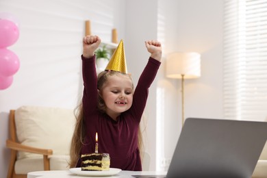 Photo of Birthday celebration. Little girl with piece of cake having video call via laptop at table indoors