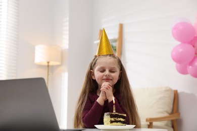 Photo of Birthday celebration. Little girl with piece of cake having video call via laptop at table indoors