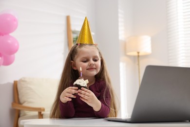 Photo of Birthday celebration. Little girl with cupcake having video call via laptop at table indoors