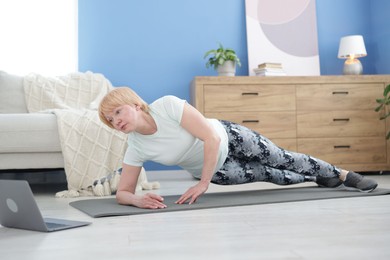 Photo of Senior woman in sportswear doing plank exercise during online class with coach at home
