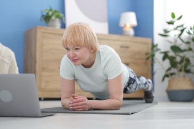 Photo of Senior woman in sportswear doing plank exercise during online class with coach at home