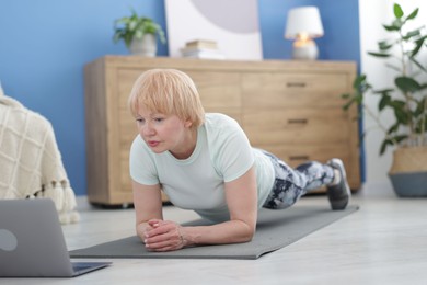 Photo of Senior woman in sportswear doing plank exercise during online class with coach at home