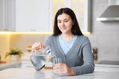 Photo of Woman pouring water into glass at white marble table in kitchen