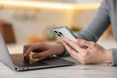 Photo of Woman using modern smartphone and laptop at white marble table indoors, closeup