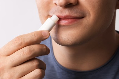 Photo of Young man applying lip balm onto his lips on light grey background, closeup
