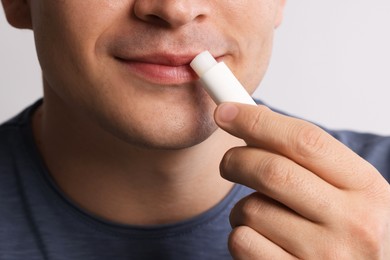 Photo of Young man applying lip balm onto his lips on light grey background, closeup