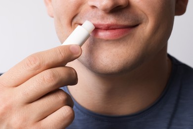 Photo of Young man applying lip balm onto his lips on light grey background, closeup
