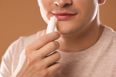 Photo of Young man applying lip balm onto his lips on light brown background, closeup