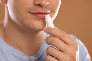 Photo of Young man applying lip balm onto his lips on light brown background, closeup