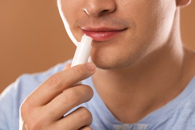 Photo of Young man applying lip balm onto his lips on light brown background, closeup