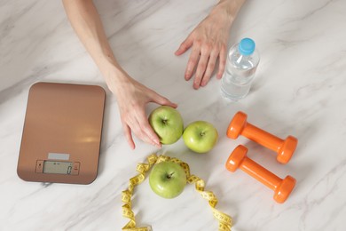 Photo of Woman at white marble table with apples, kitchen scale, dumbbells, water and measuring tape, top view
