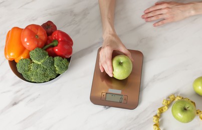 Photo of Woman weighting apple on kitchen scale at white marble table, top view