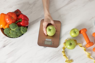 Photo of Woman weighting apple on kitchen scale at white marble table, top view