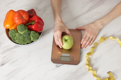 Photo of Woman weighting apple on kitchen scale at white marble table, top view