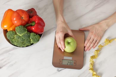 Photo of Woman weighting apple on kitchen scale at white marble table, top view
