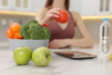 Photo of Woman weighting tomato on kitchen scale at white marble table indoors, selective focus