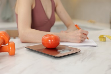 Photo of Woman developing diet plan at white marble table indoors, focus on kitchen scale and tomato
