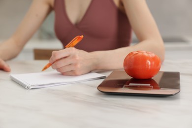 Photo of Woman developing diet plan at white marble table indoors, focus on kitchen scale and tomato