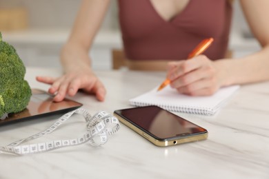 Photo of Woman developing diet plan while weighting broccoli on kitchen scale at white marble table indoors, closeup