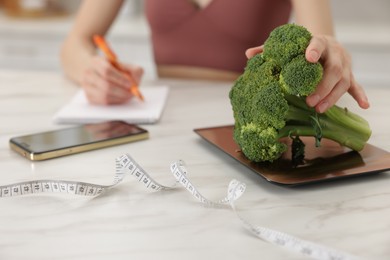 Photo of Woman developing diet plan while weighting broccoli on kitchen scale at white marble table indoors, closeup