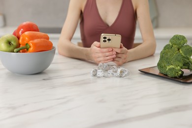 Photo of Woman using smartphone at white marble table with vegetables and measuring tape indoors, closeup