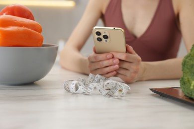 Photo of Woman using smartphone at white marble table with vegetables, kitchen scale and measuring tape indoors, closeup