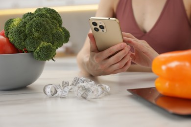Photo of Woman using smartphone at white marble table with vegetables, kitchen scale and measuring tape indoors, closeup