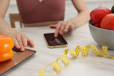 Photo of Woman using smartphone while weighting bell pepper on kitchen scale at white marble table indoors, closeup